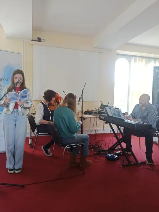 A young writer reading at the microphone, with a violinist and a keyboard player