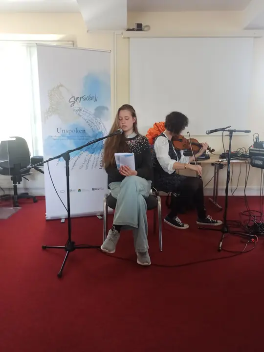 A young writer reading seated at the microphone while a violinist plays