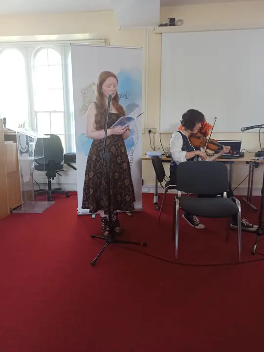 A young writer reading at the microphone while a violinist plays